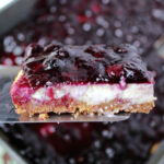 Easy blueberry cheesecake bars on a spatula with a pan behind.