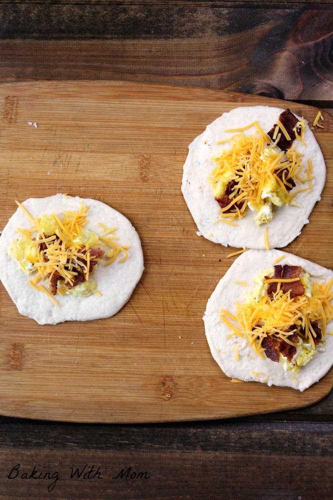 Biscuits on a cutting board, flattened, with bacon and cheese on top. 