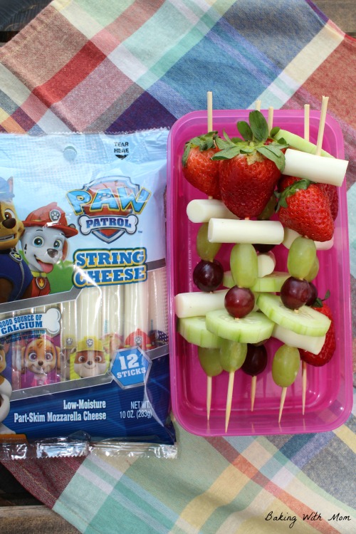 Fruit and cheese kabobs in a container with a package of string cheese laying besides. 