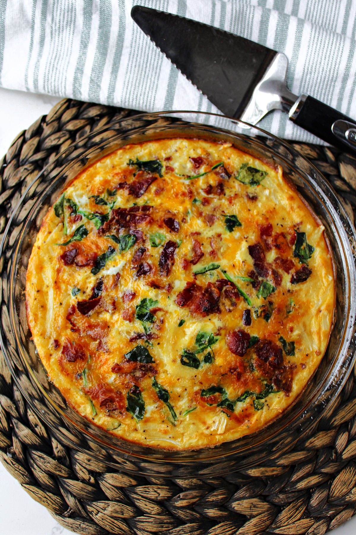 bacon spinach quiche on a brown mat with a pie server nearby. 