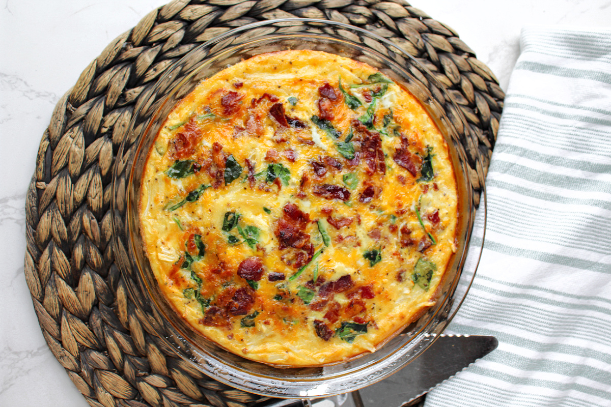 bacon spinach quiche in a pie pan with a towel nearby.