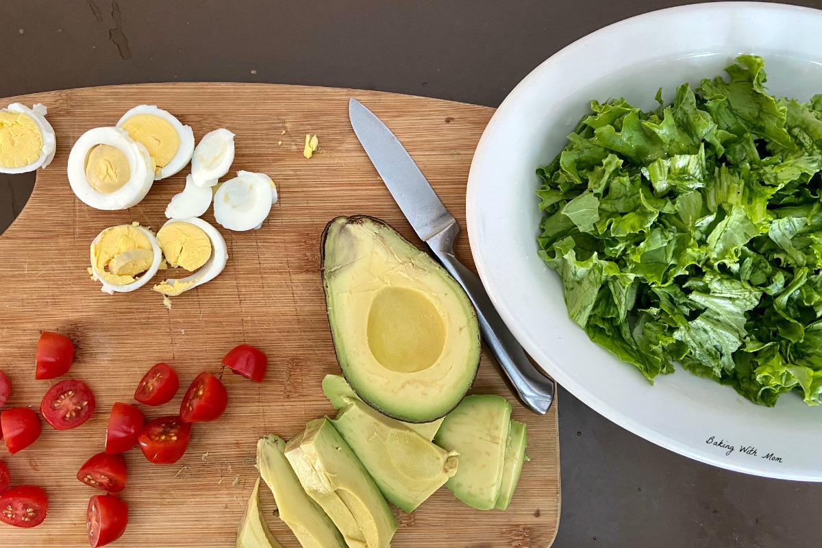 Cutting board with avocado, tomatoes, hard boiled eggs and leafy lettuce in a bowl.  