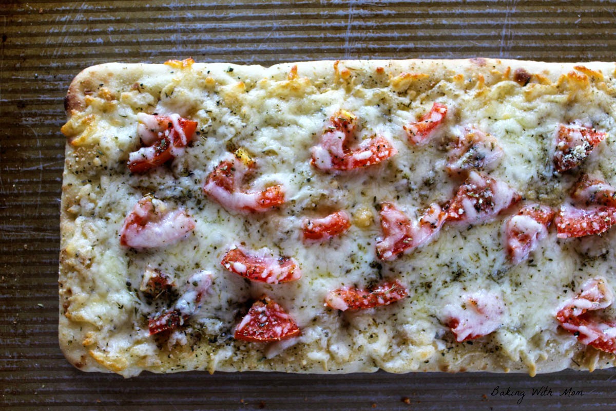 Flatbread on a baking sheet with layers of cheese, tomatoes, Italian seasoning