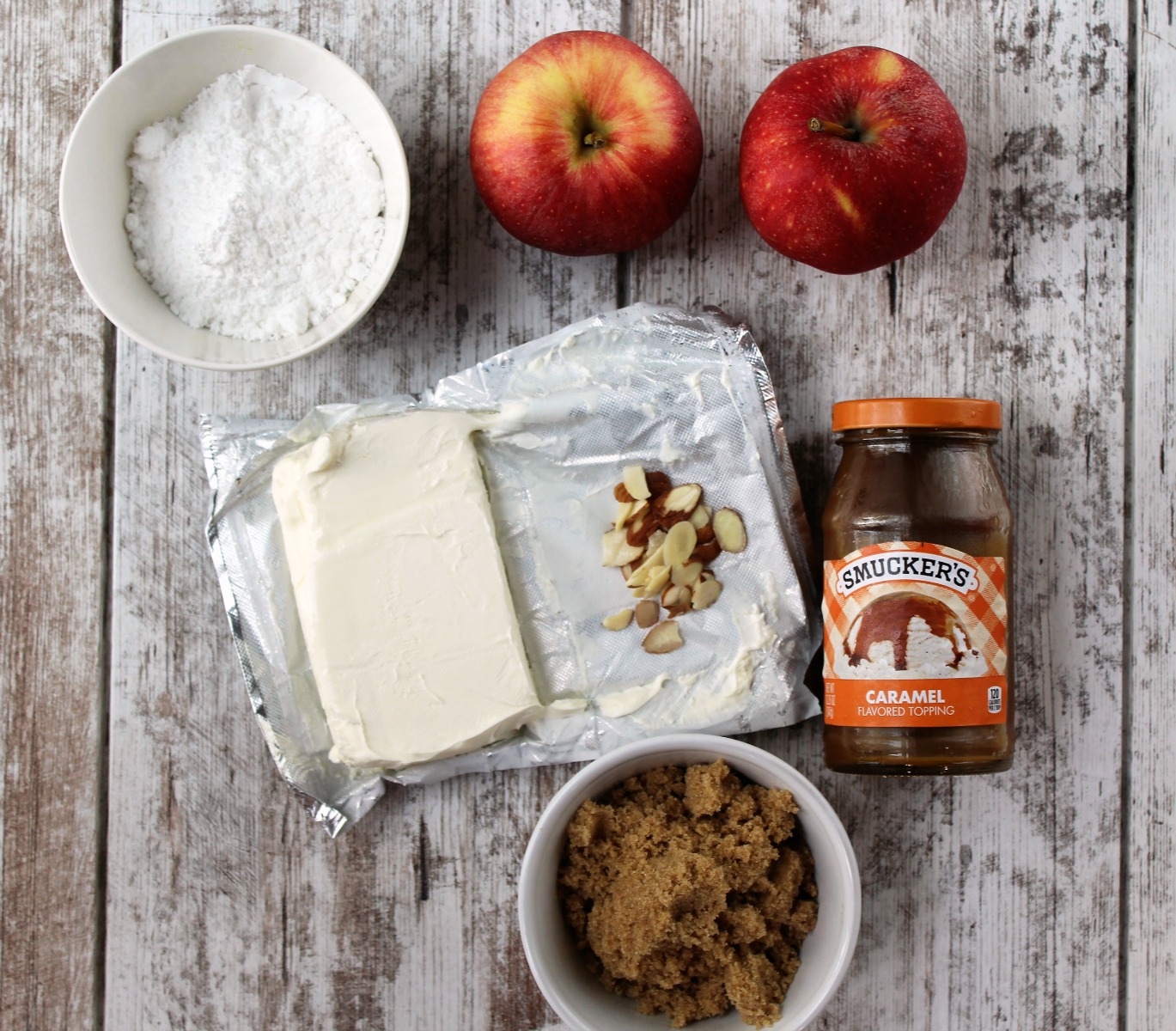 Powdered sugar, red apples, jar of caramel sauce, block of cream cheese, brown sugar and almonds