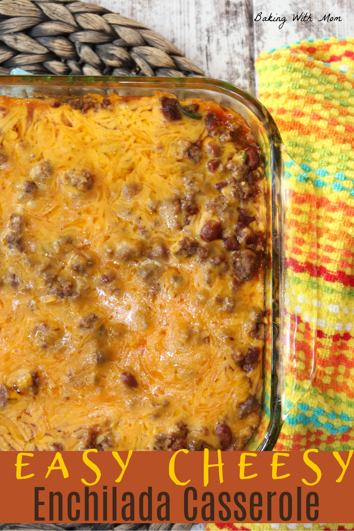 Enchilada casserole in a casserole dish with a colorful towel next to it. Layers of cheese, beans and hamburger.