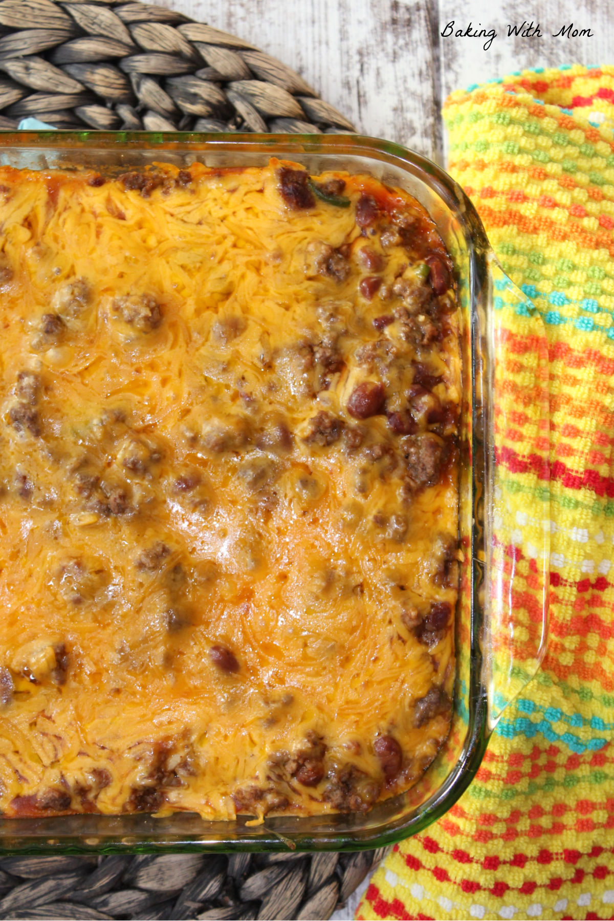 Enchilada casserole with cheddar cheese on top in a clear casserole dish with a multi-colored towel lying nearby. 