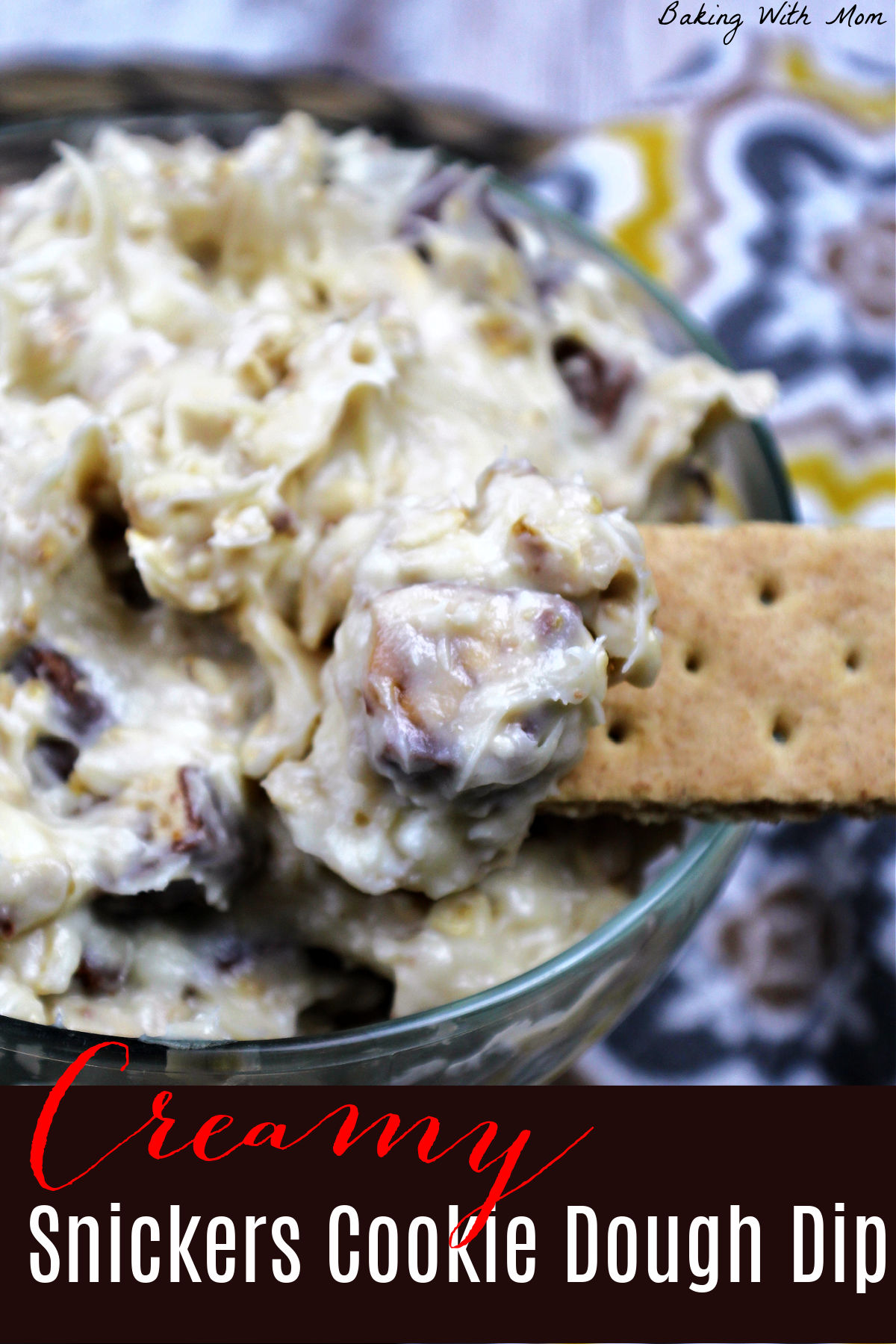 Cookie dough dip with snickers in a clear bowl with a yellow and gray towel laying besides.