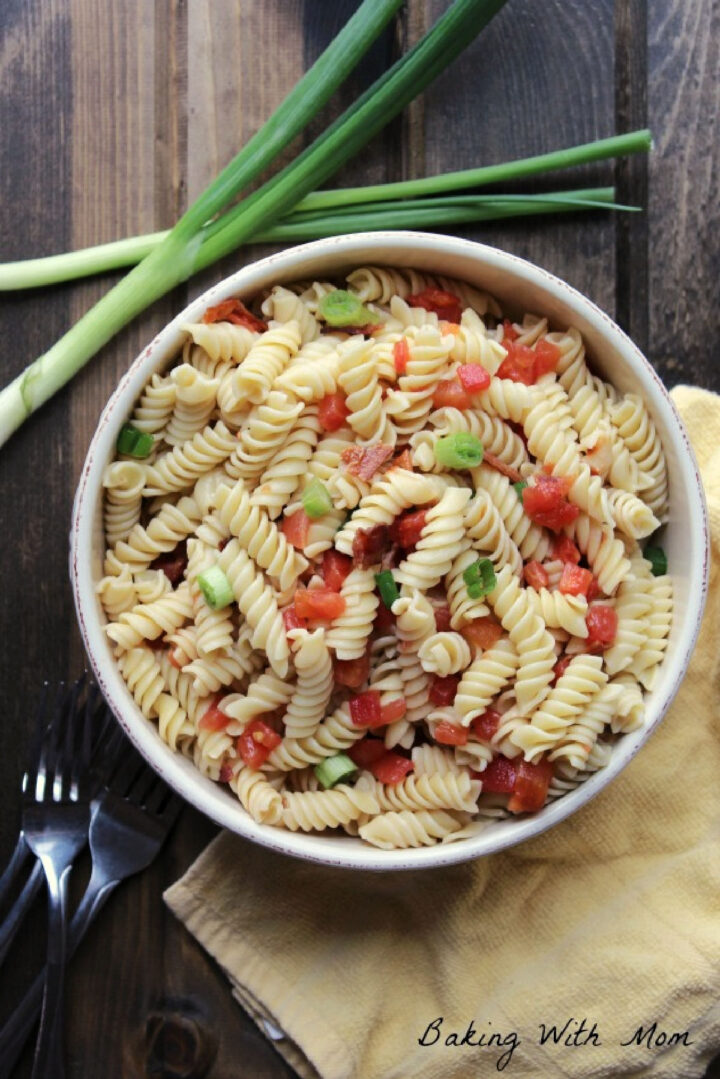 Pasta salad recipe with green onions and tomatoes in a cream colored bowl with green onions laying besides.