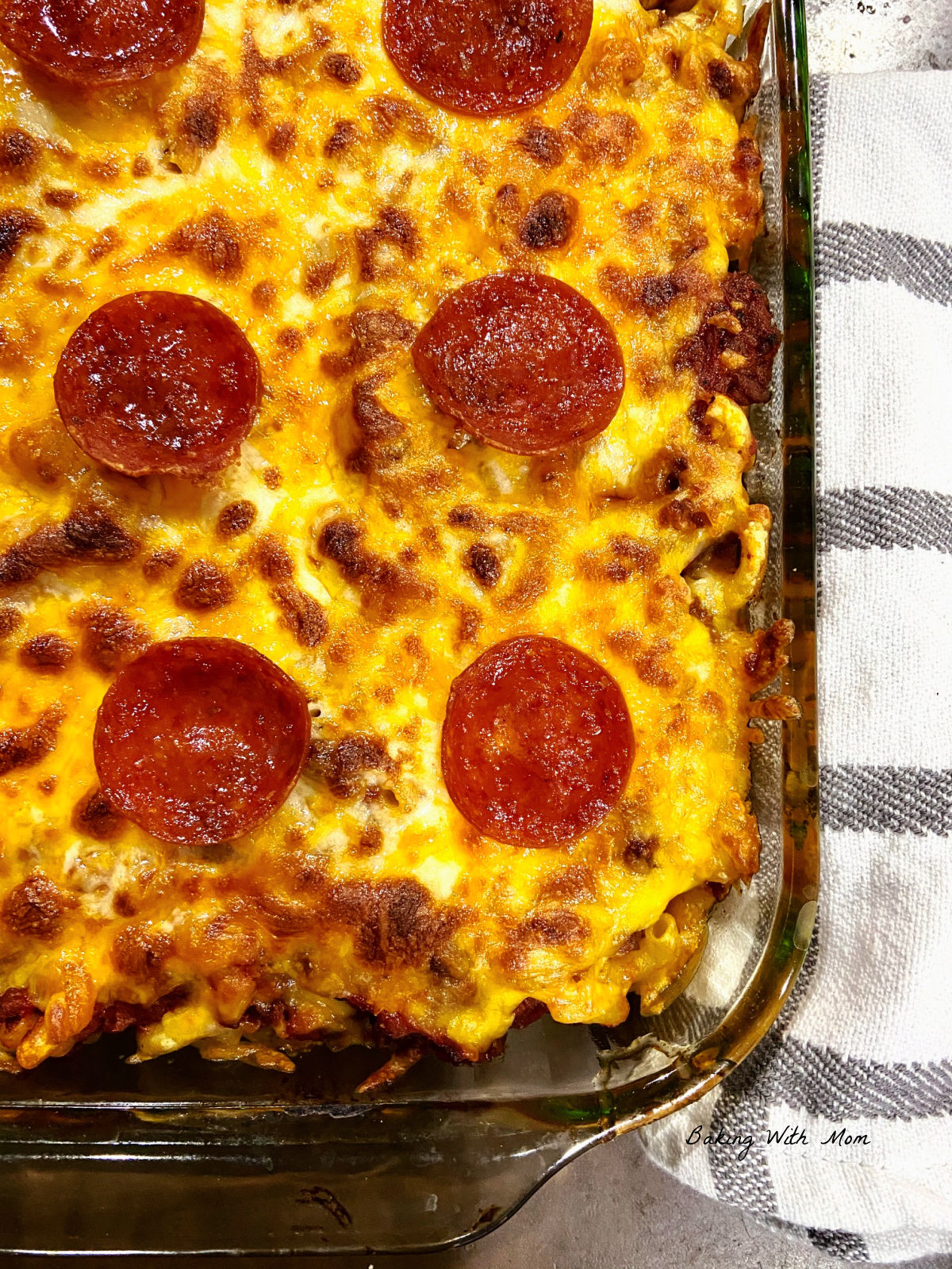 Casserole dish with noodles, cheese and pepperoni on top. A towel is laying besides. 