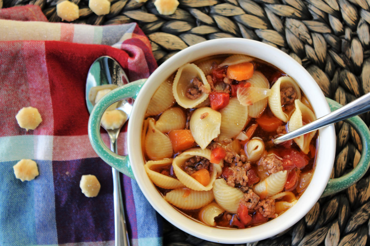 Bowl of hamburger soup with shells in a blue bowl with tomatoes and carrots. 