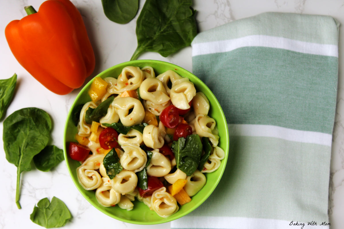 Bowl of tortellini salad with a towel, orange pepper and spinach laying besides.