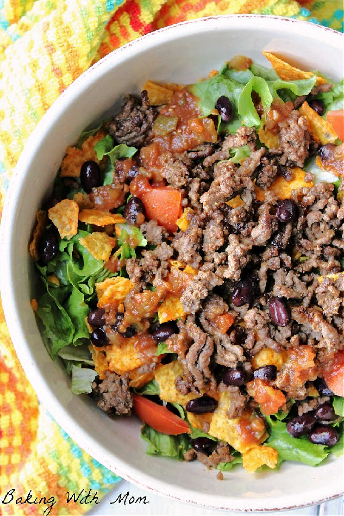 Taco salad in a white bowl white bowl with a multicolored towel laying besides. 