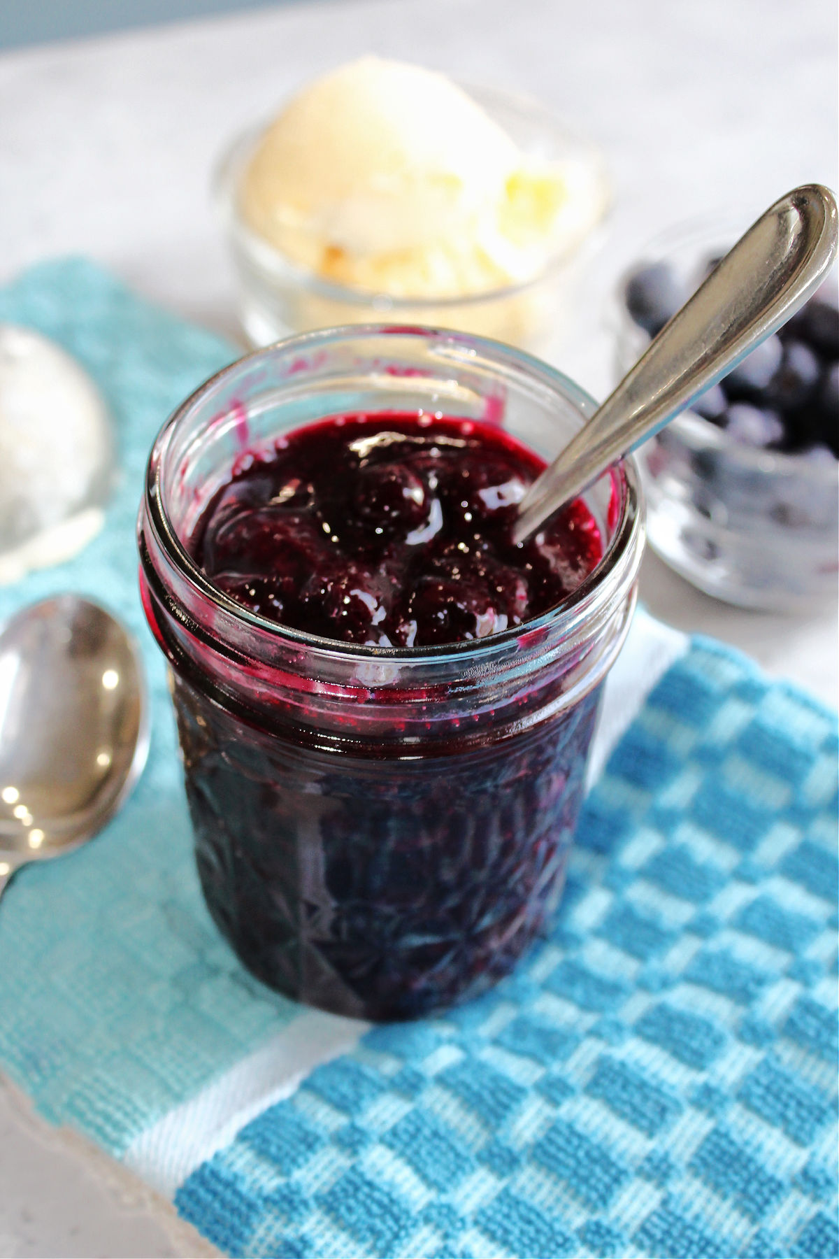 jar of homemade blueberry sauce on a blue towel with ice cream in the background. 