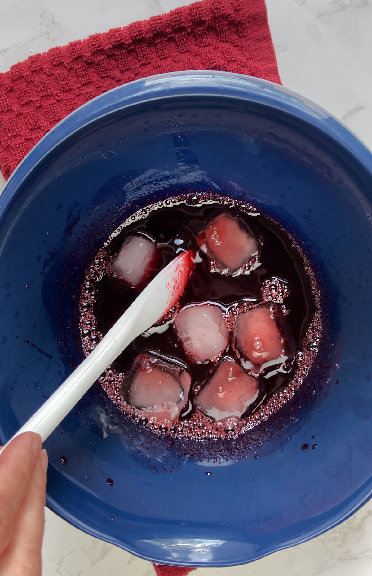 Hand stirring ice cubes and strawberry gelatin in a blue bowl. 