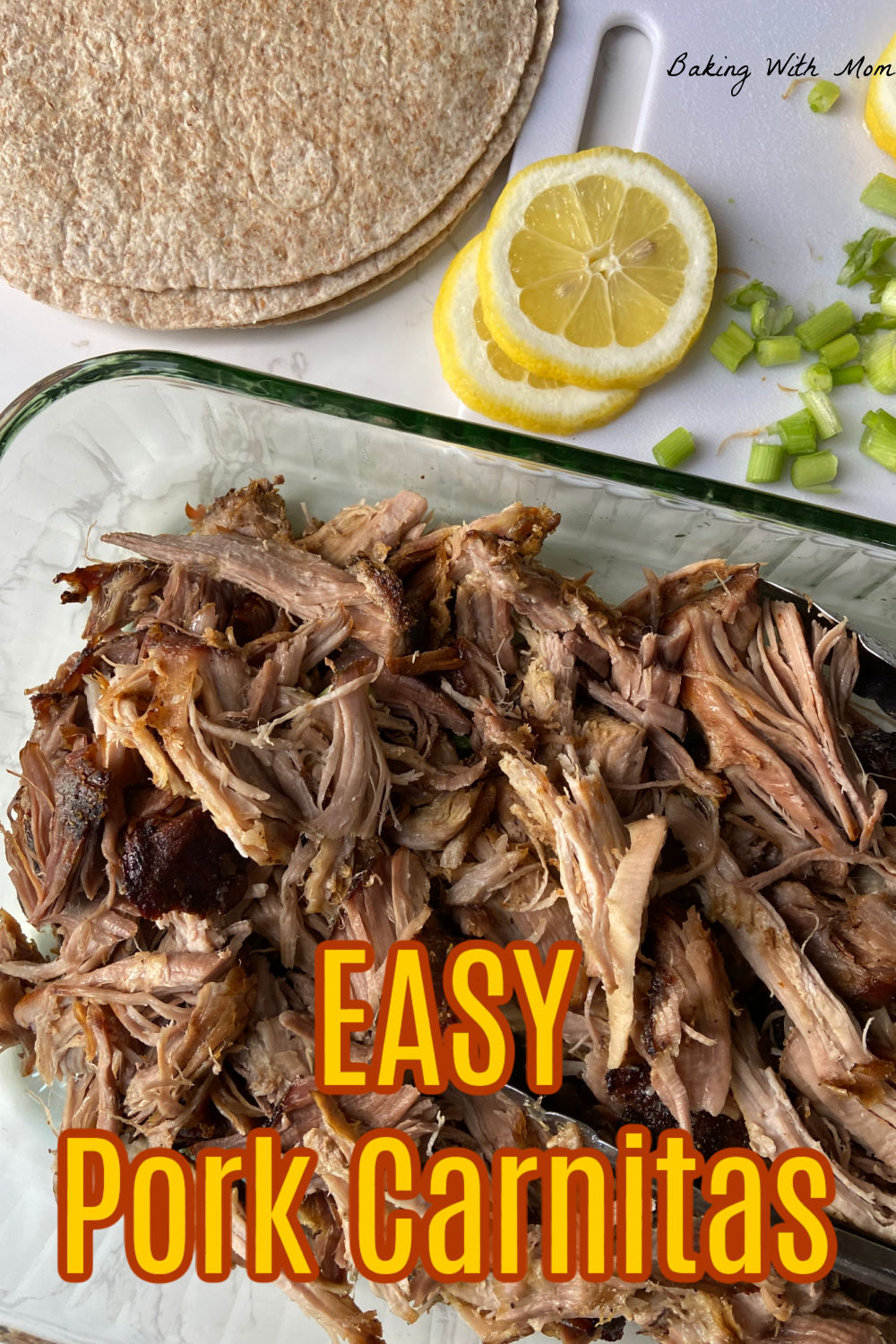 picture of pork carnitas in a baking dish with tortillas, green onions and lemons nearby. 