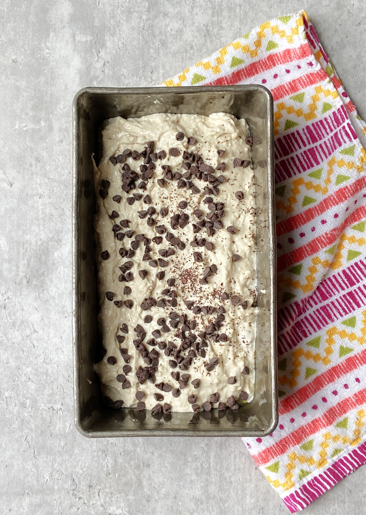 Chocolate chips on top of unbaked bread in a loaf pan with a towel lying besides. 