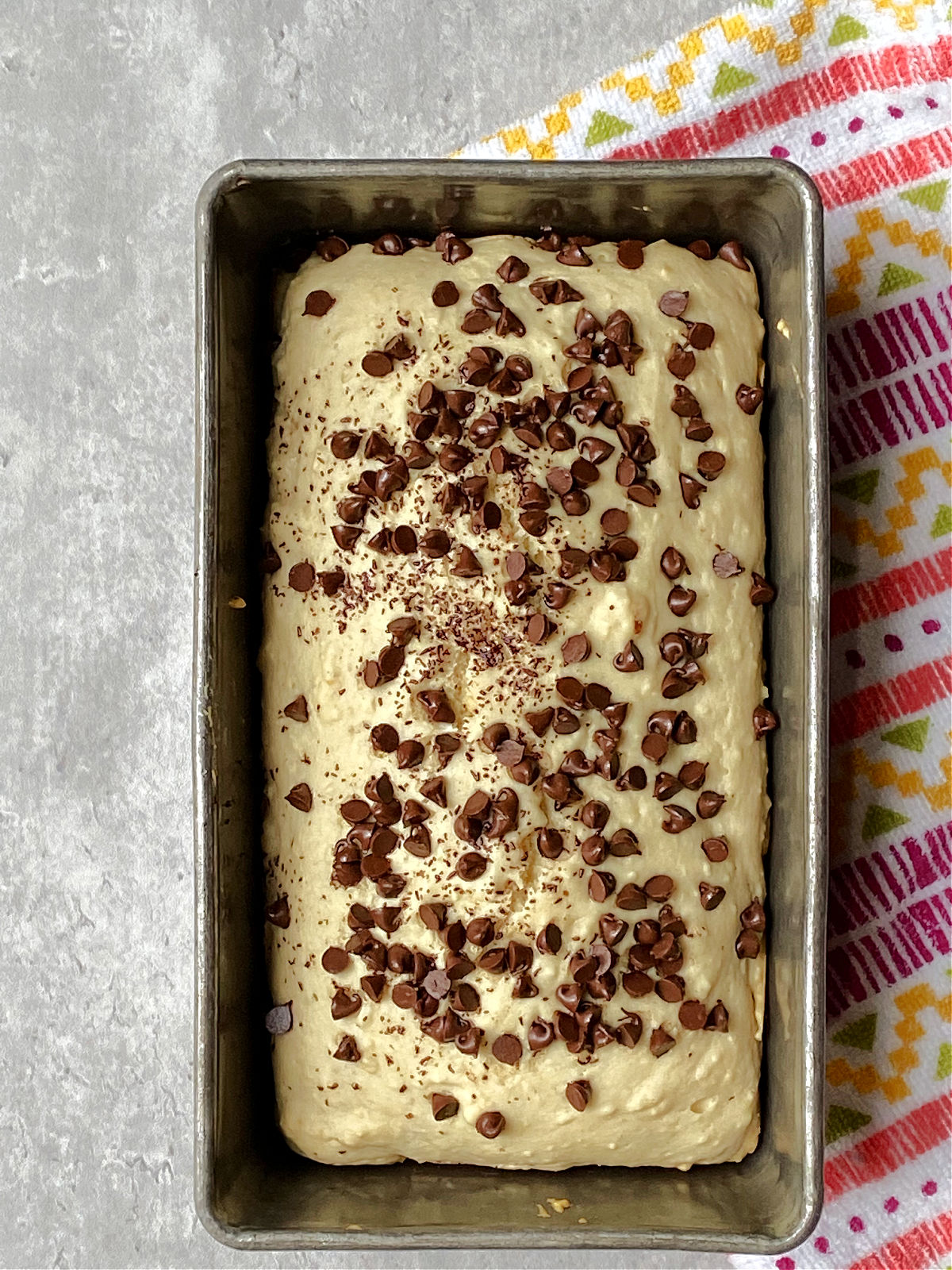 Chocolate chip ice cream bread in a loaf pan with a multicolored towel lying besides. 