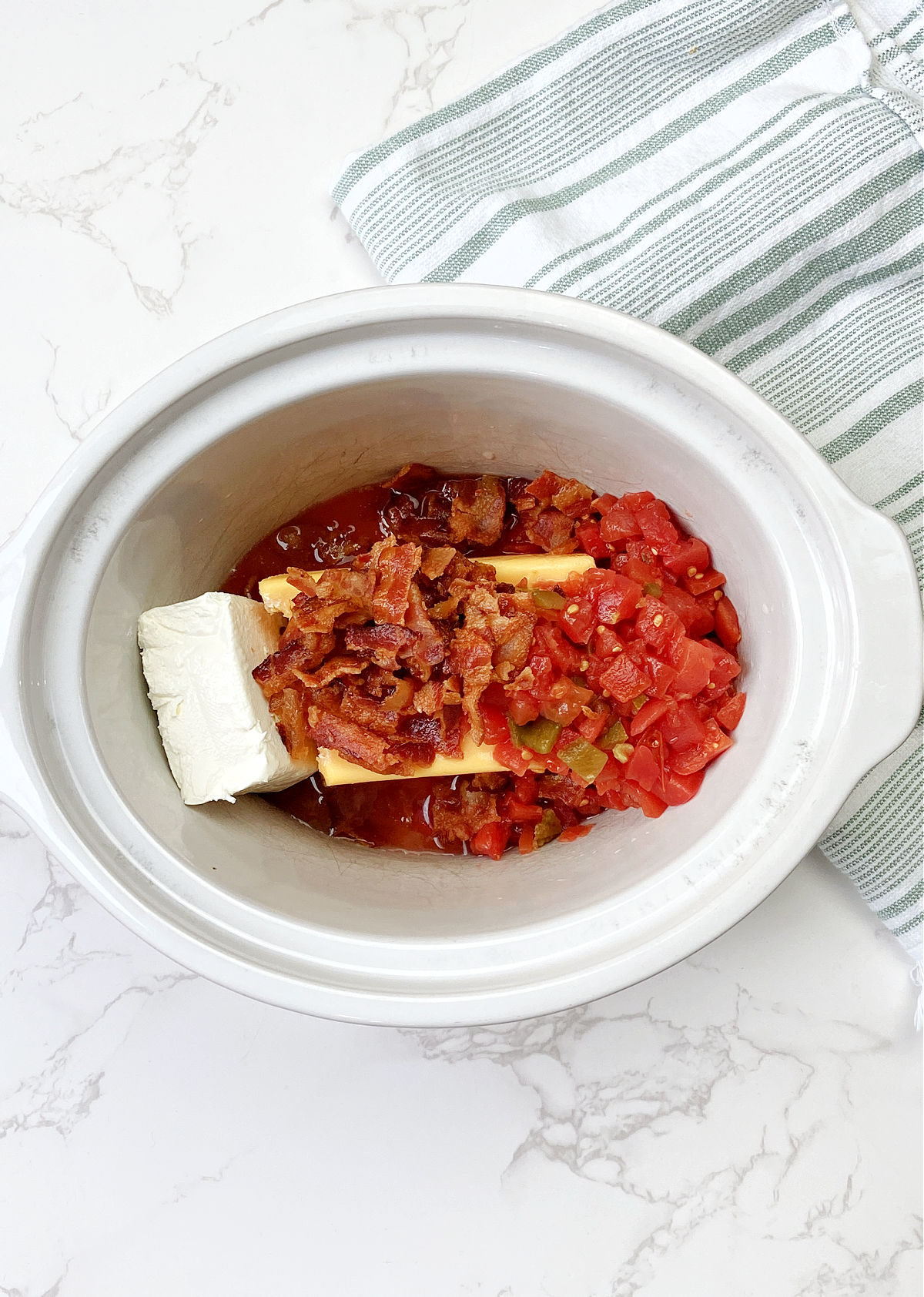 Bacon on top of Velveeta cheese with cream cheese, tomatoes in a white crock pot. 