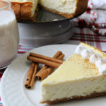 Eggnog cheesecake on a white plate with cinnamon sticks besides.