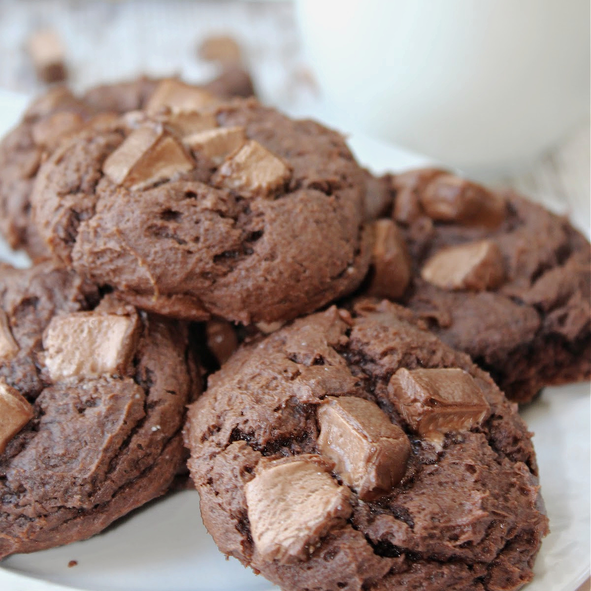 Gooey chocolate cookies on a white plate with milk behind them.